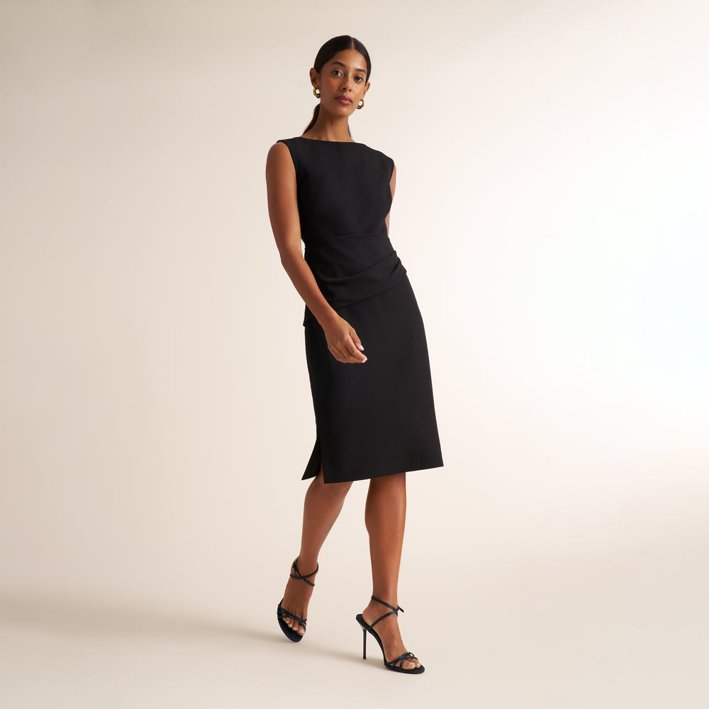 A woman modeling a sleeveless black knee-length dress with a structured boat neckline and a draped waist detail, paired with black strappy high-heeled sandals against a warm off-white studio background.