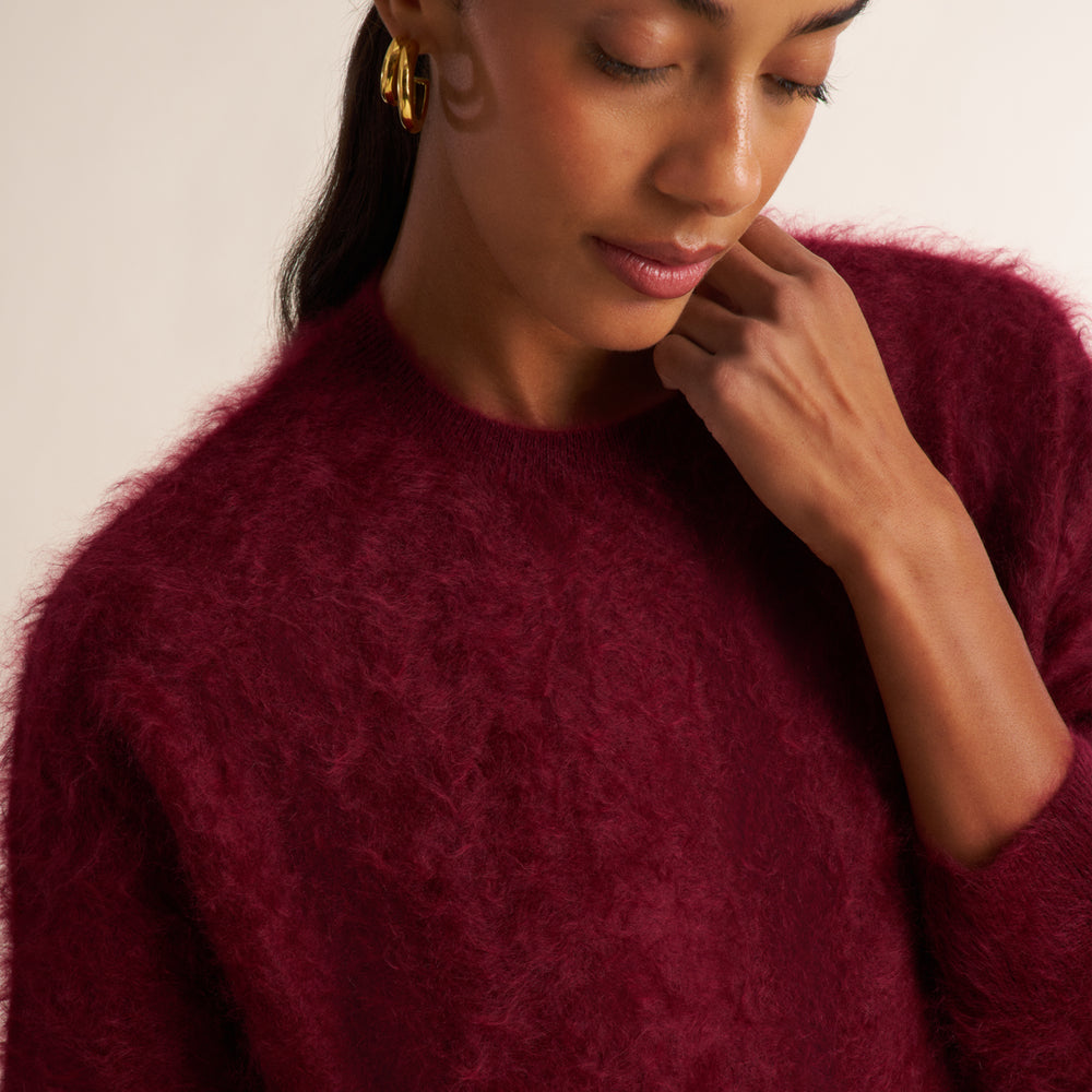 A close-up of a woman wearing a fuzzy, mohair-like crewneck sweater in deep burgundy. She is wearing gold hoop earrings and looking down, set against a plain, warm off-white background.