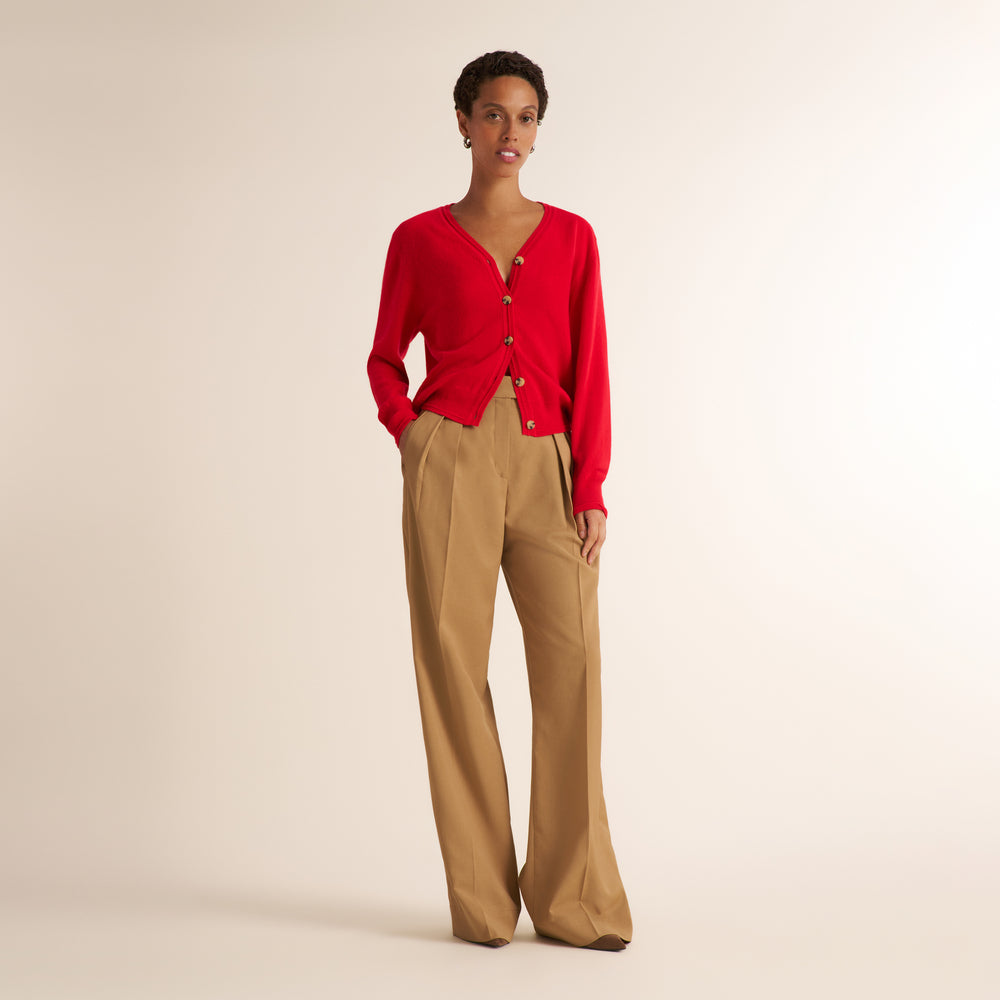 A model stands against a warm beige studio background wearing a bright red wrap-style cardigan with buttons and tan wide-leg trousers.