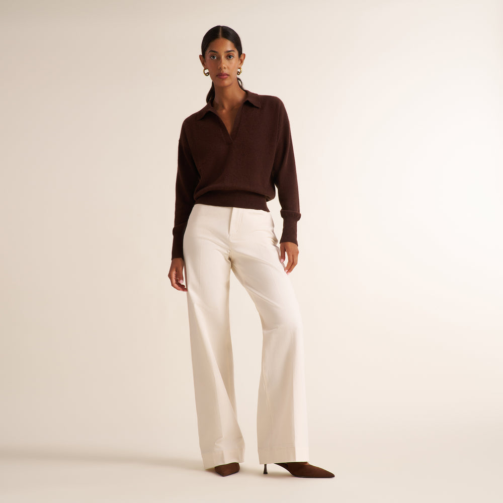 A model stands against a warm off-white studio background wearing a dark brown long-sleeve collared knit top, off-white wide-leg trousers, and dark brown pointed-toe heels.