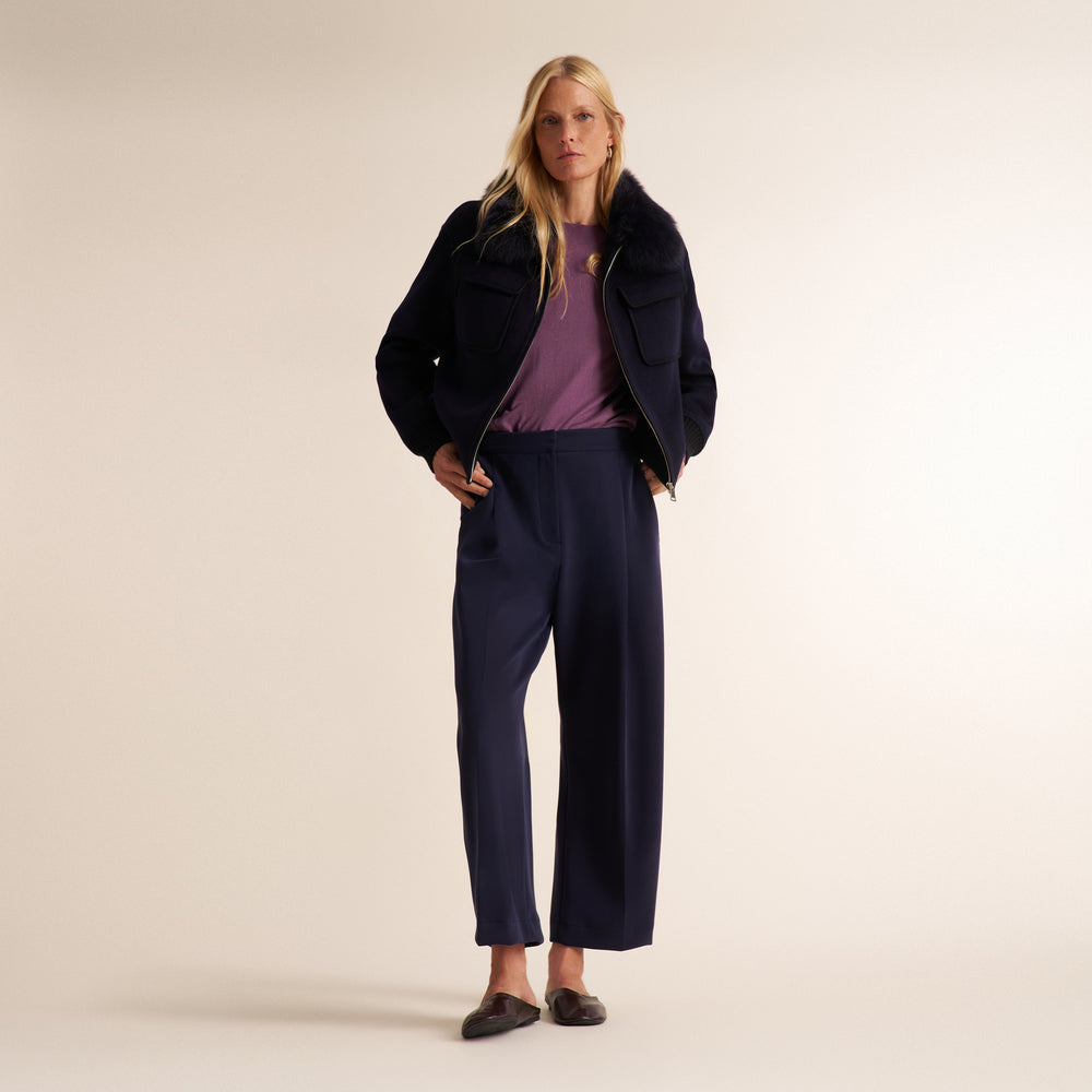 A woman stands against a plain warm beige studio background wearing a dark navy cropped jacket over a mauve-purple top and matching navy wide-leg trousers, with dark flats.