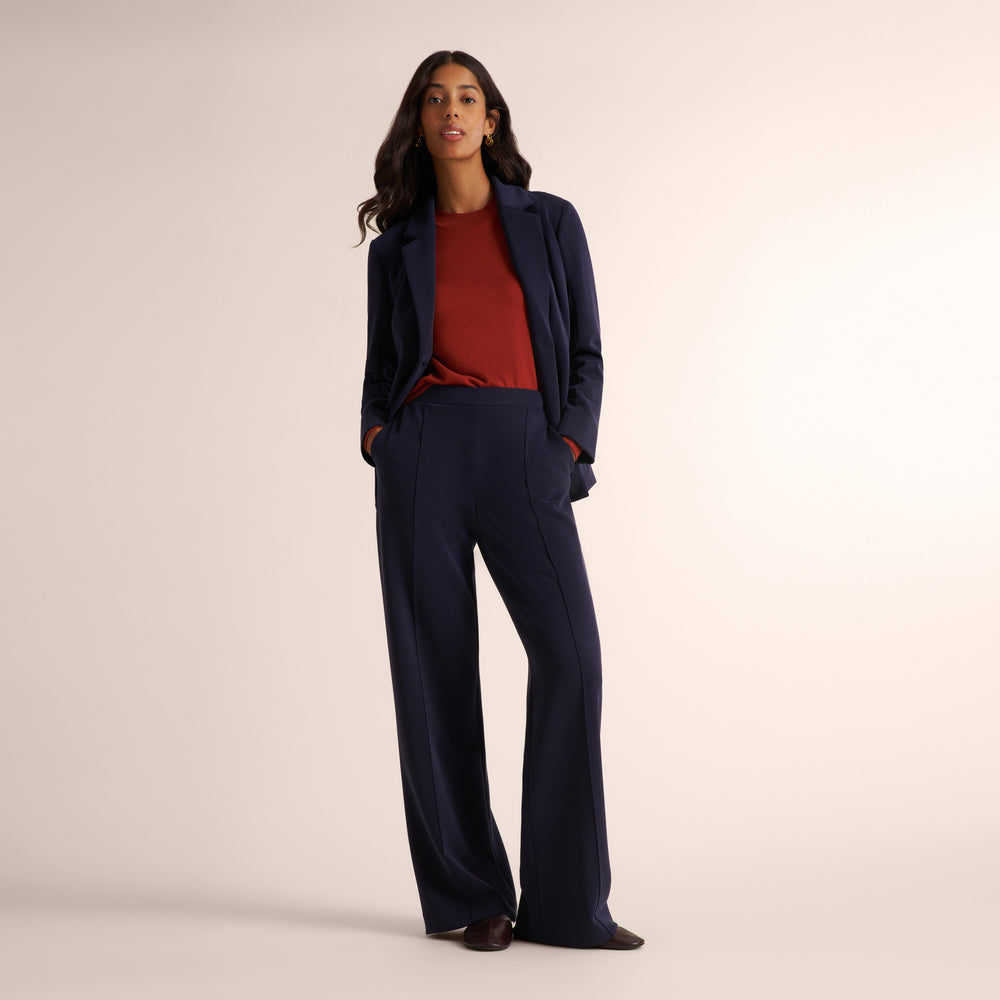 A woman modeling a navy blue wide-leg blazer and matching trousers, styled with a rust-red top, standing against a solid light beige studio background.