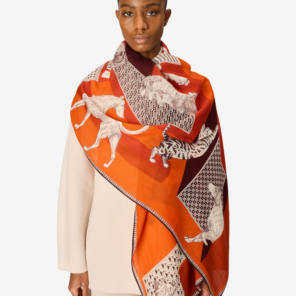 A model wearing a vibrant orange and red patterned silk scarf with white and black animal prints, styled over a cream-colored top against a light gray background.