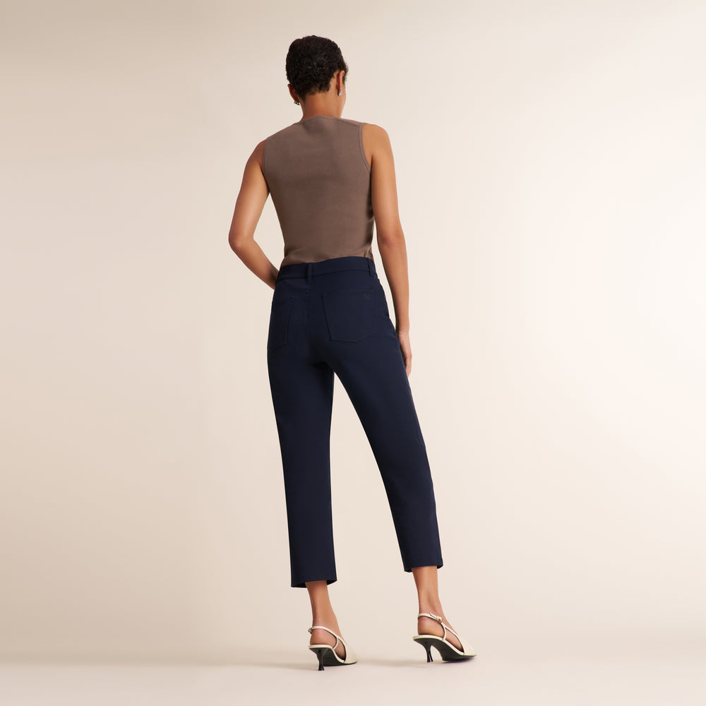 A woman shown from the back wearing navy blue cropped trousers, a taupe sleeveless top, and strappy high-heeled sandals against a plain warm beige studio background.