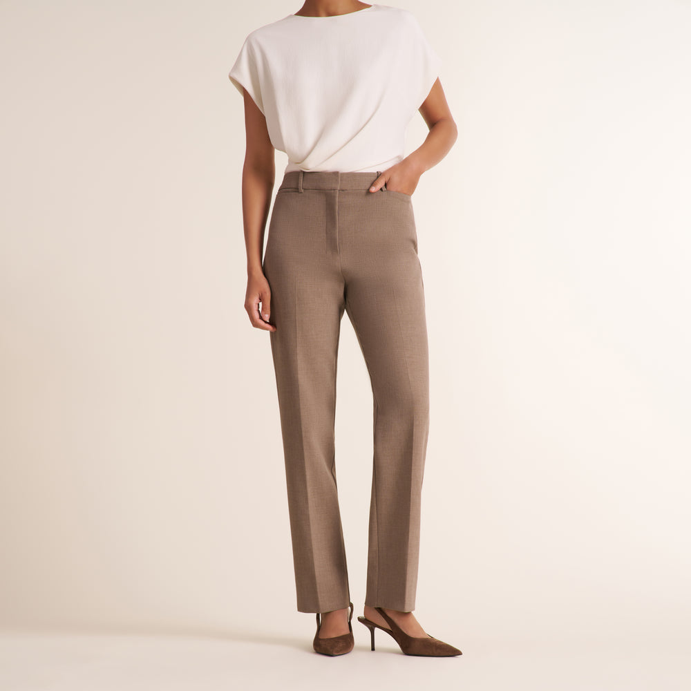 A model from the back wearing high-waisted taupe straight-leg trousers, a white draped short-sleeve top, and brown pointed-toe slingback heels against a warm off-white studio background.