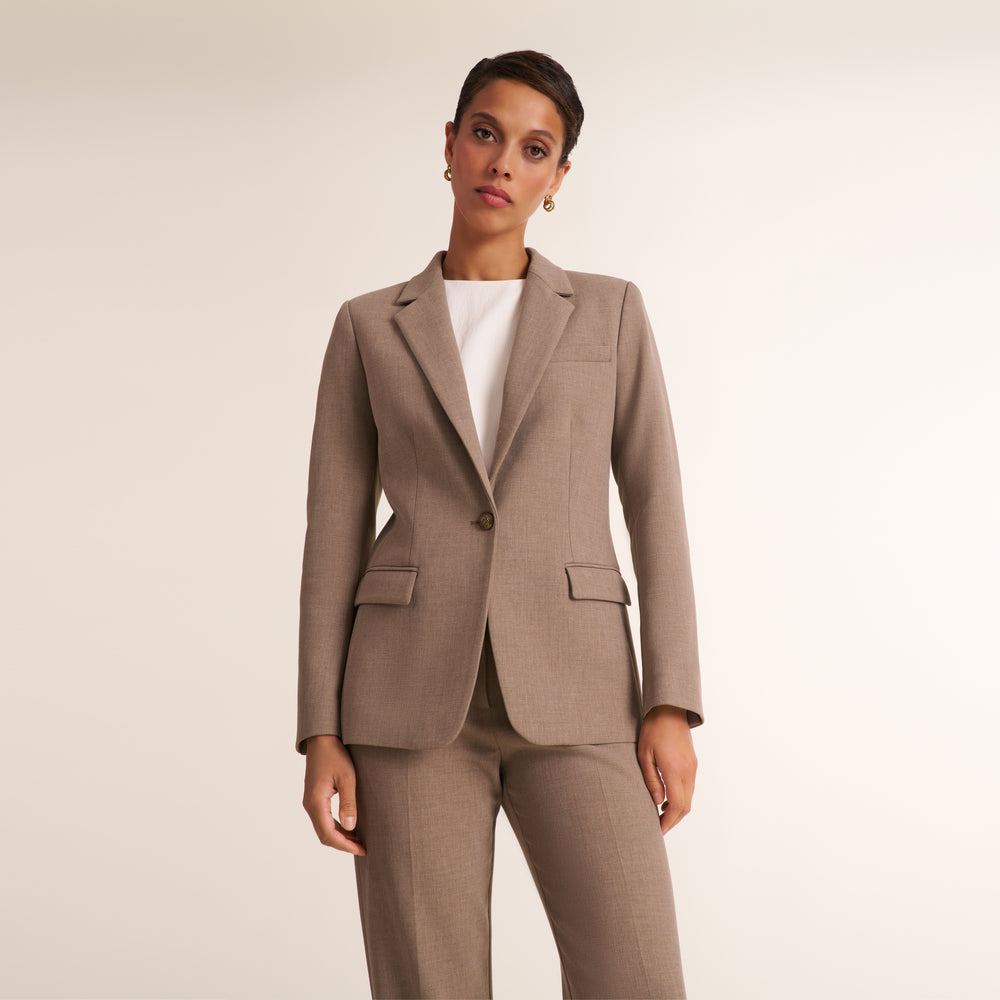 A woman modeling a tailored taupe blazer with matching trousers over a white top, photographed against a plain light beige studio background.