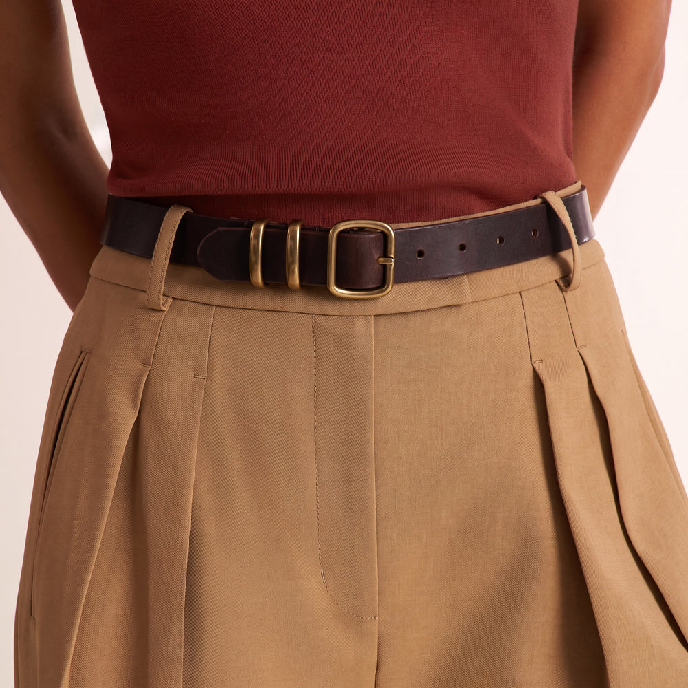 A close-up of a person wearing a dark brown leather belt with a gold-tone buckle, styled with pleated tan trousers and a rust-red ribbed top against a light beige background.