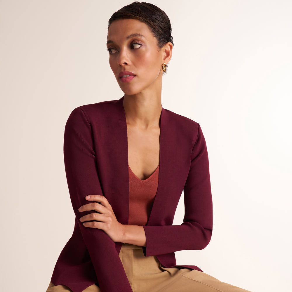 A woman wearing a deep burgundy open-front blazer over a rust-colored top and beige trousers, posed against a clean off-white studio background.