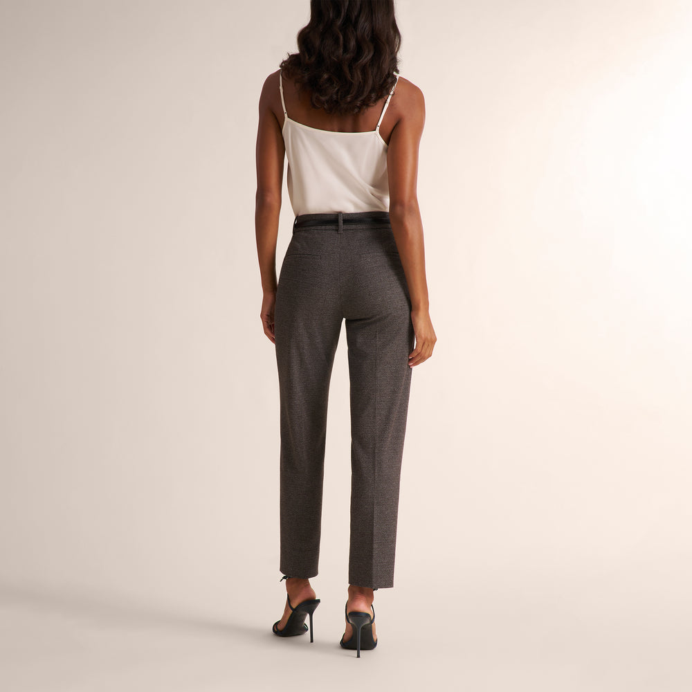 A woman is shown from the back wearing high-waisted, straight-leg charcoal gray trousers with a thin black belt, paired with a white camisole and black heels, against a warm off-white studio background.