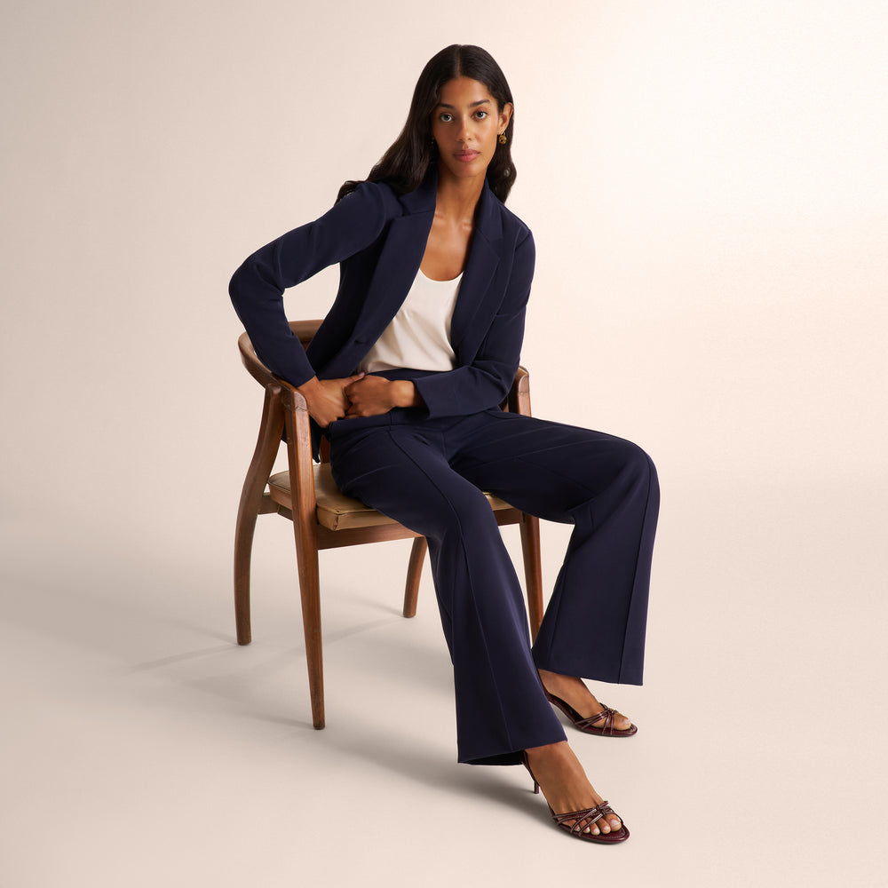 A woman sits on a wooden chair wearing a navy blue tailored blazer and matching wide-leg pants over a white top, photographed in a clean studio setting.