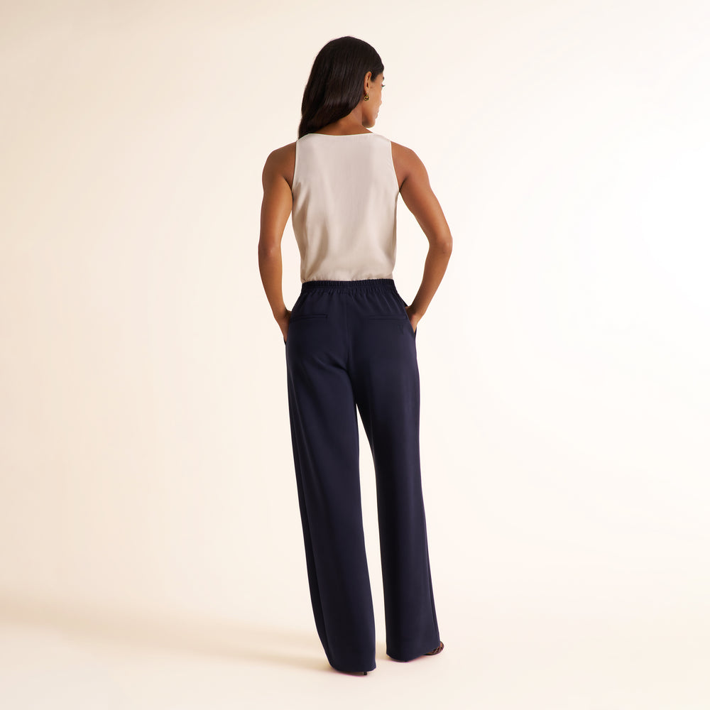A woman is shown from behind wearing a sleeveless cream top and navy wide-leg pants against a plain cream studio background.