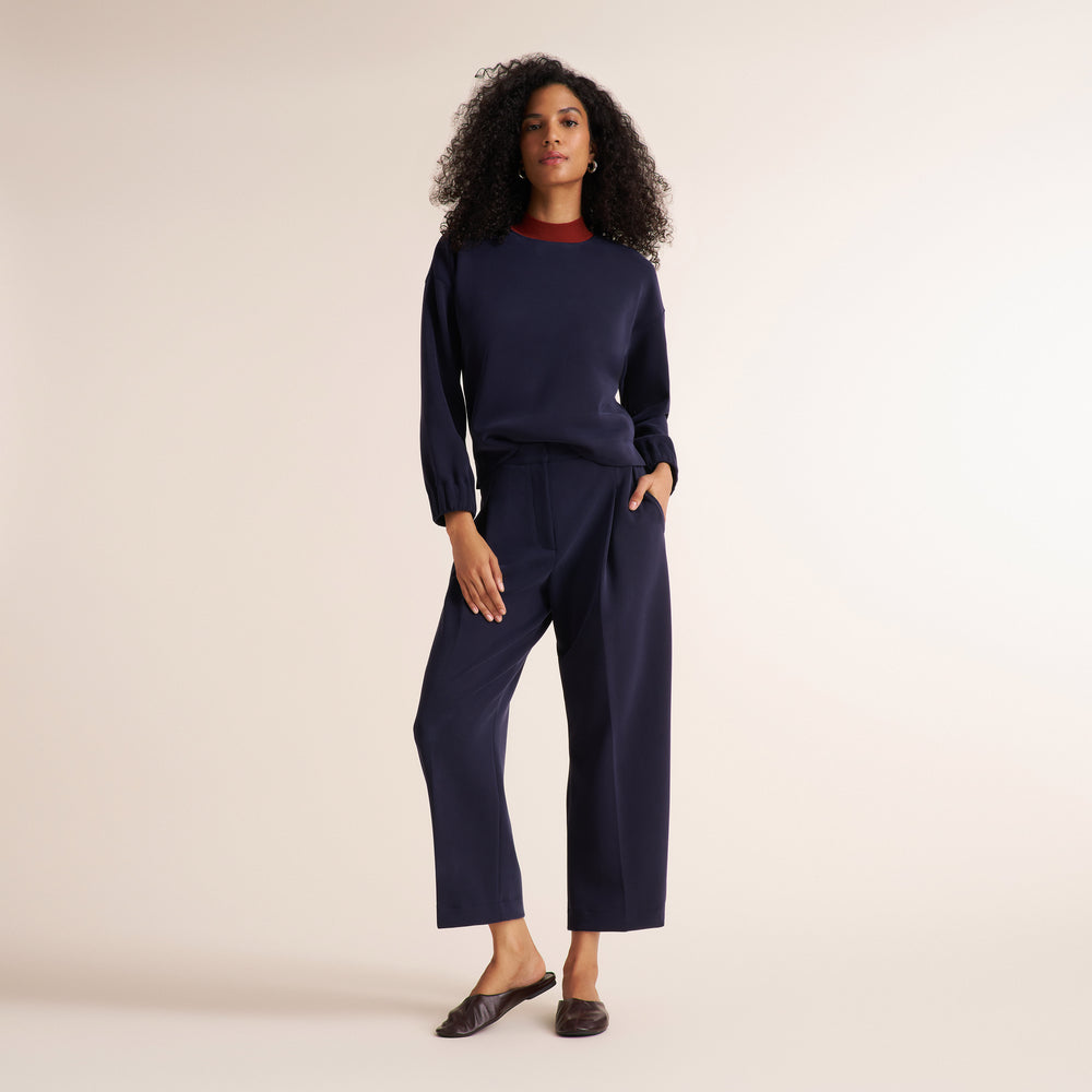 A woman modeling a navy blue knit two-piece set consisting of a long-sleeve crewneck top and matching wide-leg cropped pants, paired with dark brown flat shoes against a solid light beige studio background.