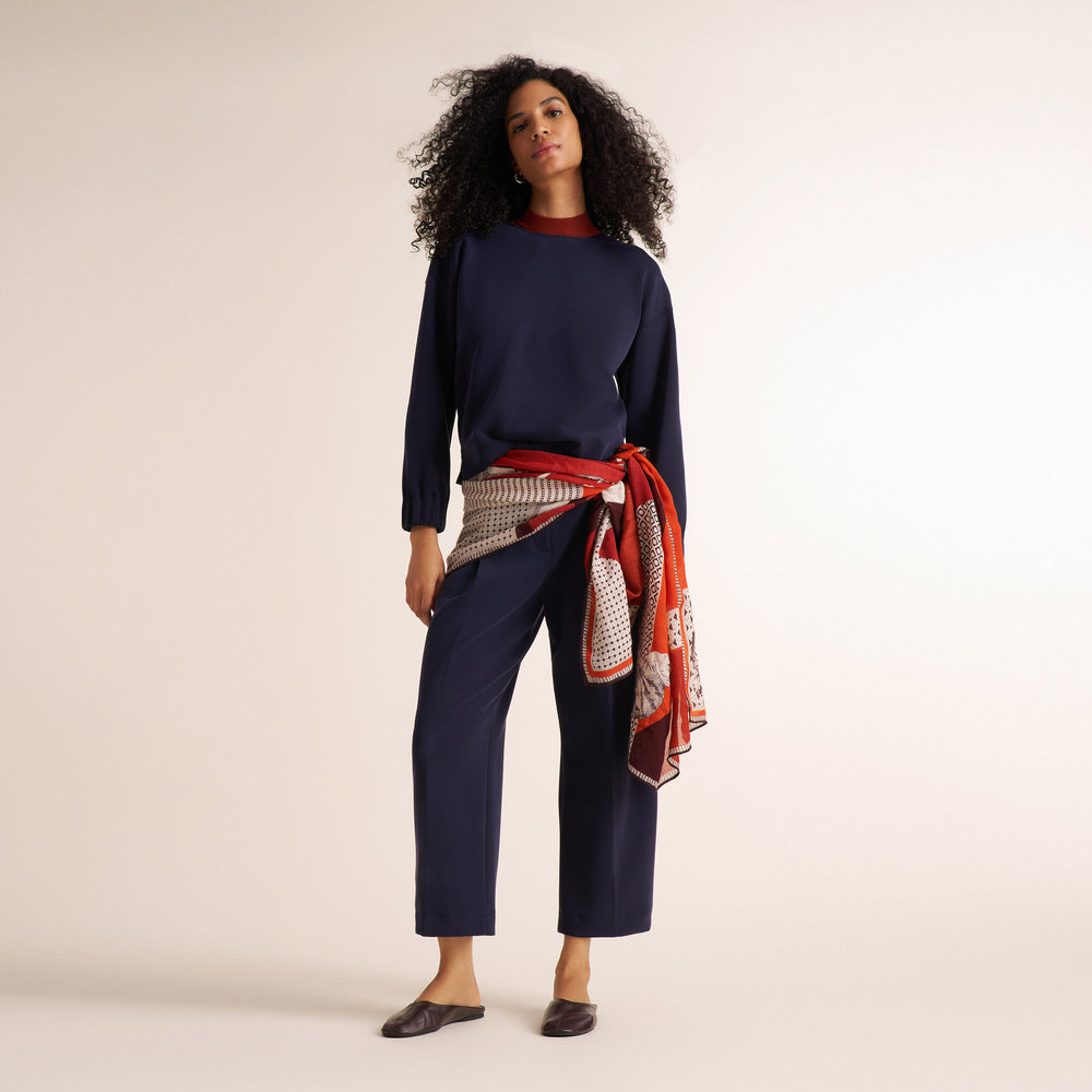 A woman stands against a plain warm beige studio background wearing a navy long-sleeve top and matching navy cropped pants, with a red undershirt peeking at the neckline and a patterned red-and-beige scarf tied around her waist. She is wearing dark flats.