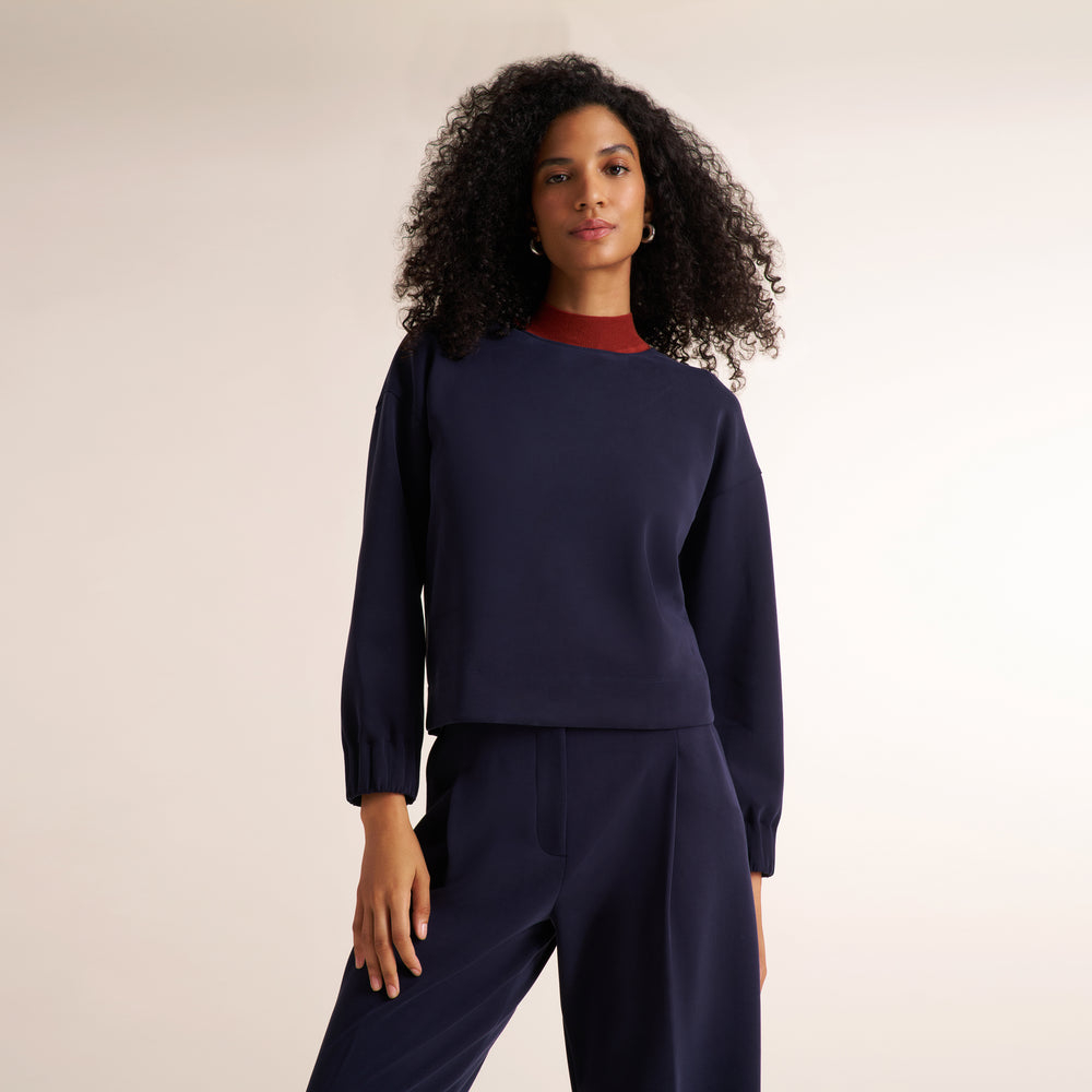 A woman with curly black hair models a navy blue long-sleeve knit top and matching navy trousers, layered over a dark red crewneck undershirt, against a plain light beige studio background.