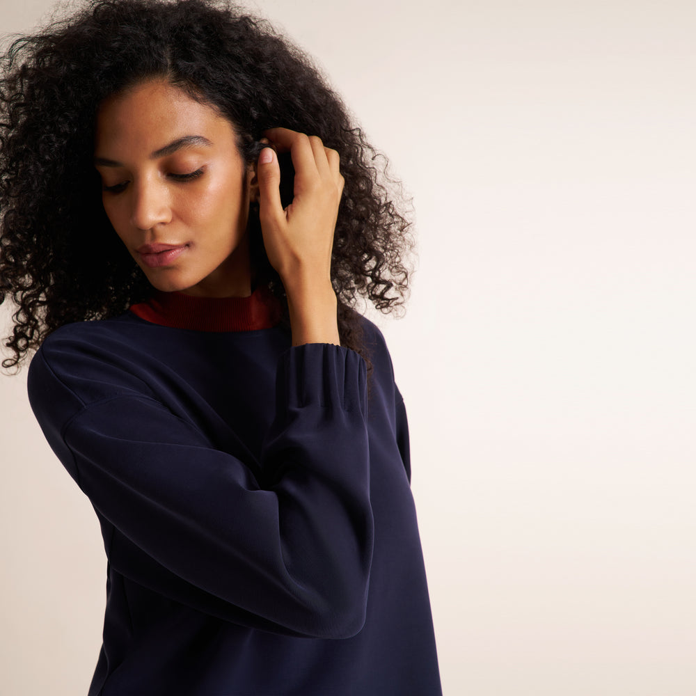 A woman with curly black hair wearing a navy blue long-sleeve top with a contrasting red mock neck collar, posing against a plain off-white background.