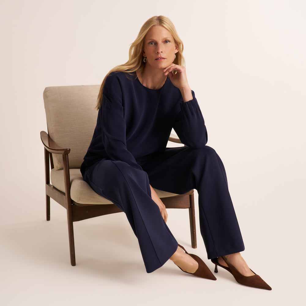 A woman sits in a beige upholstered armchair wearing a matching navy long-sleeve top and wide-leg pants with brown pointed-toe heels, photographed against a clean warm off-white studio background.