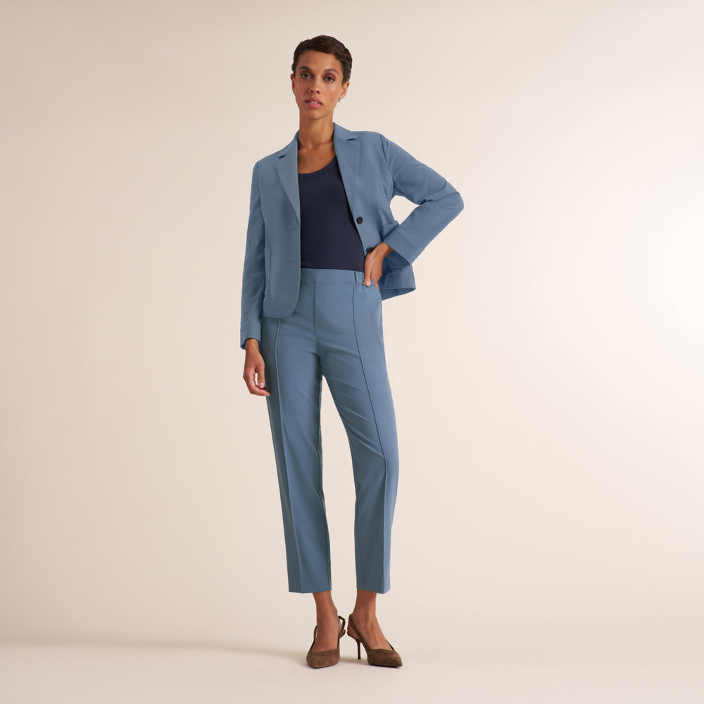 A model stands against a plain beige studio background wearing a light blue tailored suit with a blazer and cropped trousers, layered over a dark navy top and brown heels.