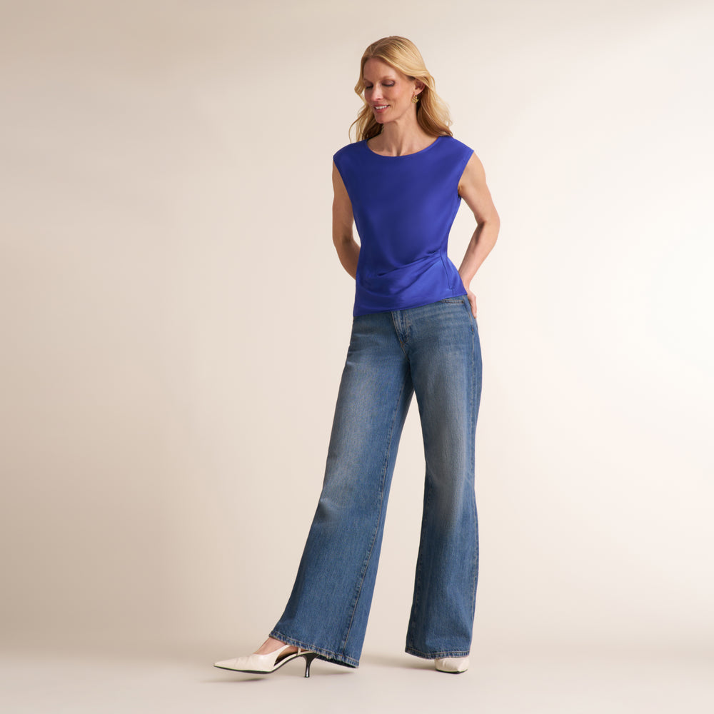 A woman modeling a sleeveless royal blue top and wide-leg blue jeans with white pointed-toe heels, standing against a plain beige studio background.