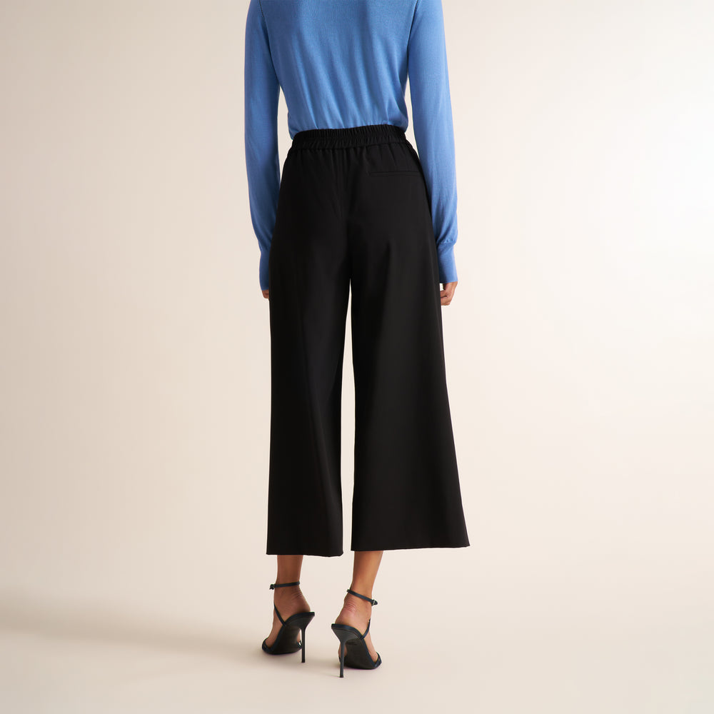 A model shown from the back wearing black wide-leg cropped pants, a blue long-sleeve top, and black high-heeled sandals against a plain beige studio background.