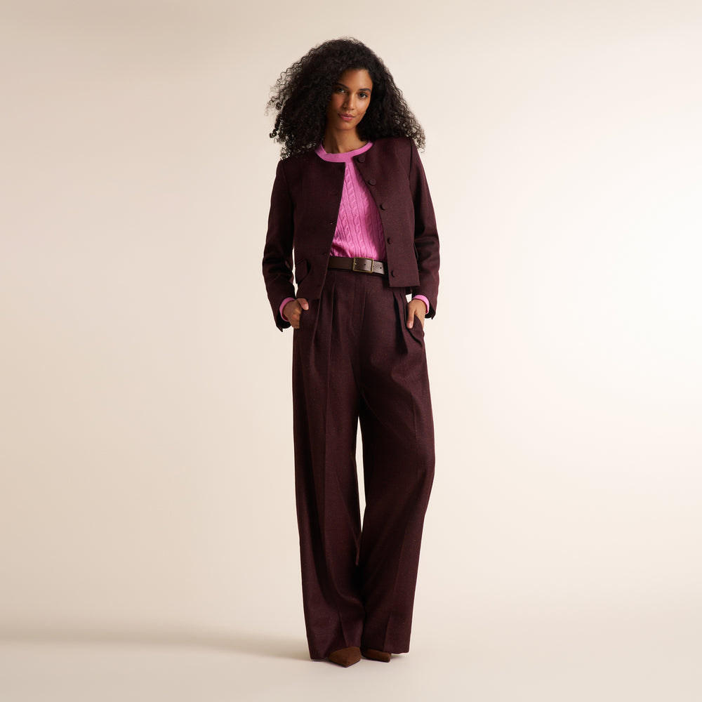 A model stands against a warm beige studio background wearing a dark burgundy two-piece suit with a pink textured top and a brown belt.