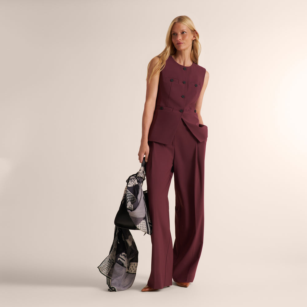 A woman models a sleeveless double-breasted jumpsuit in deep burgundy with matching wide-leg trousers and a patterned scarf, standing against a solid light beige studio background.