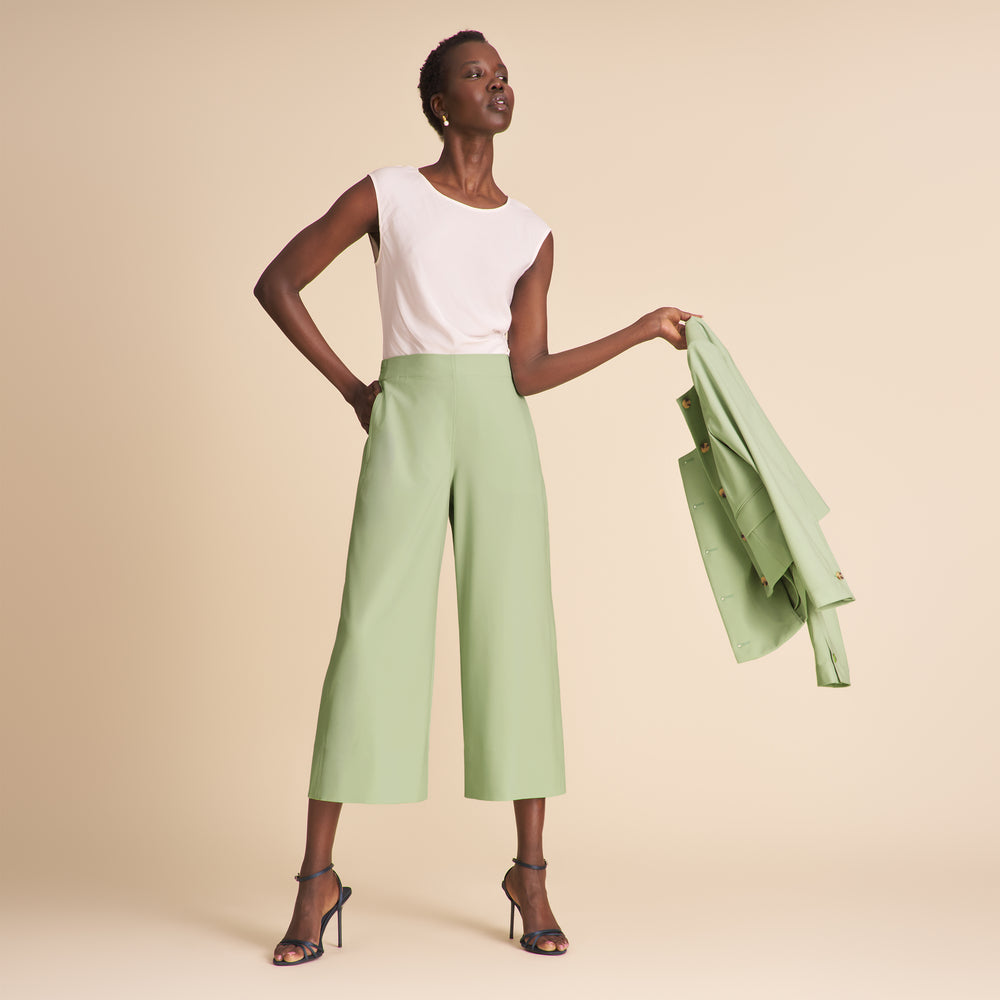 Woman wearing a white top and light green pants holding a matching jacket against a beige background