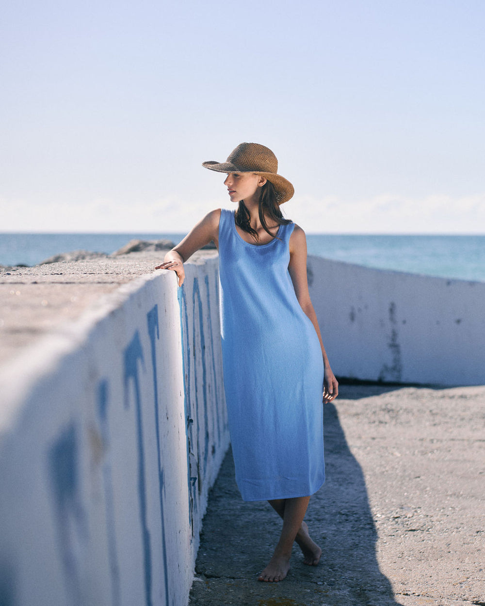 Woman in a blue dress and straw hat standing on a beach promenade.
