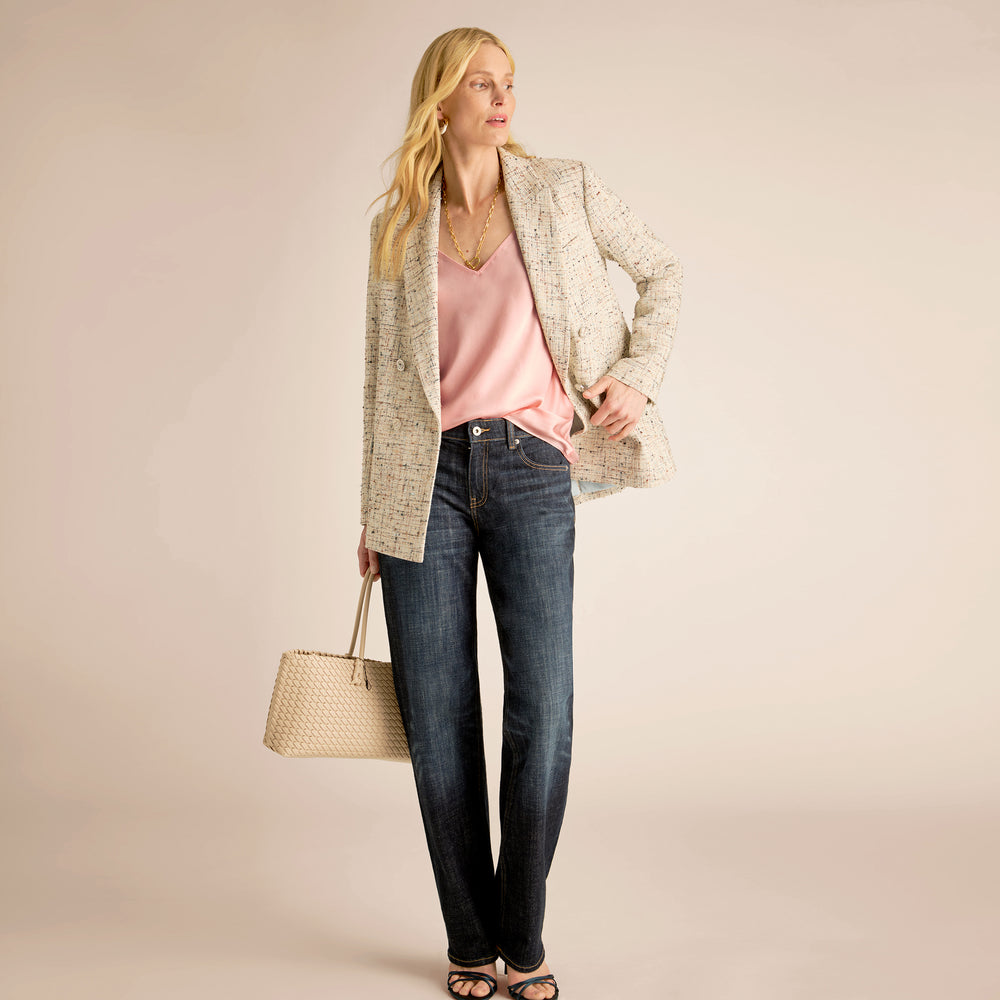 Woman wearing a light jacket, pink top, and jeans, holding a beige handbag on a beige background