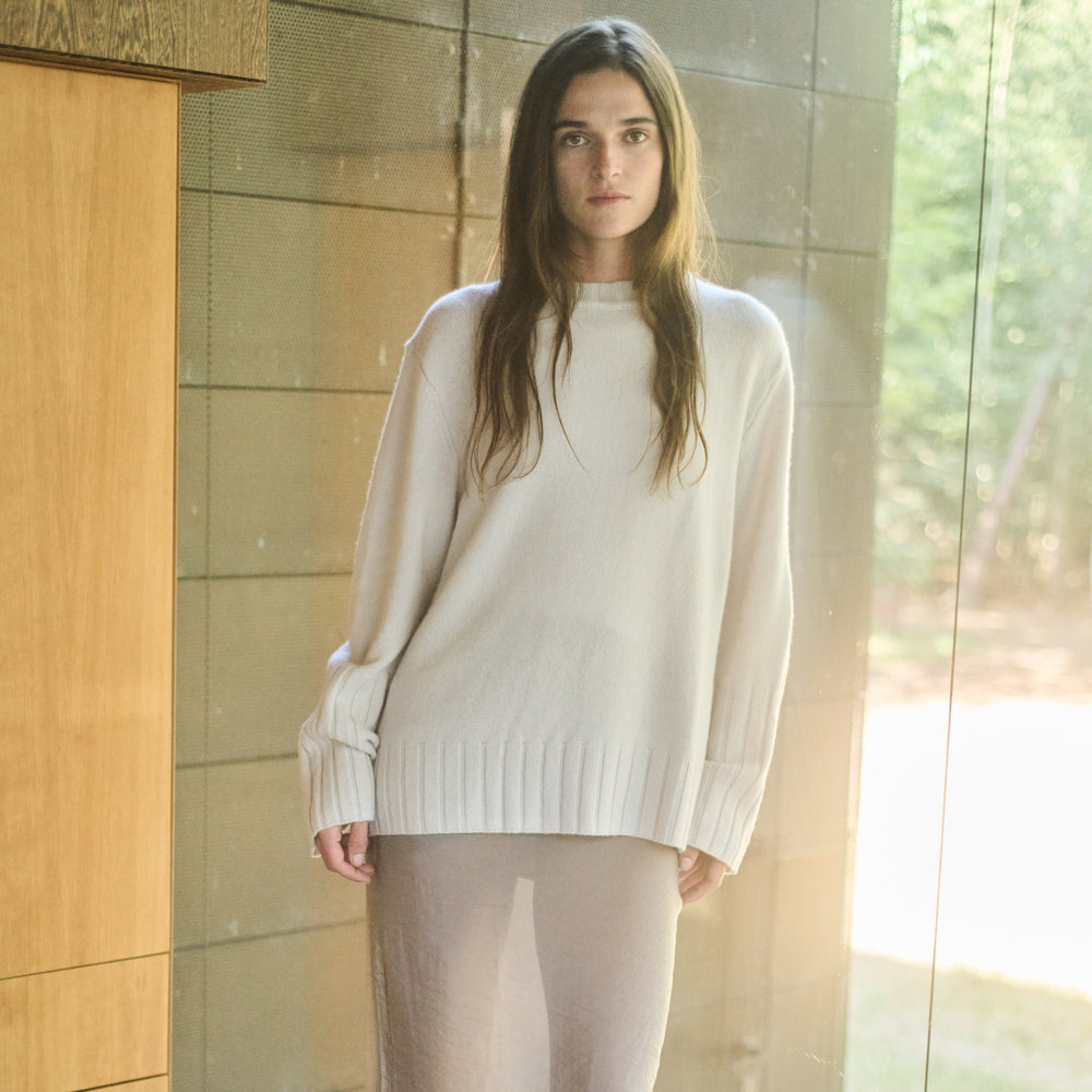Woman wearing a white sweater and beige skirt standing indoors.