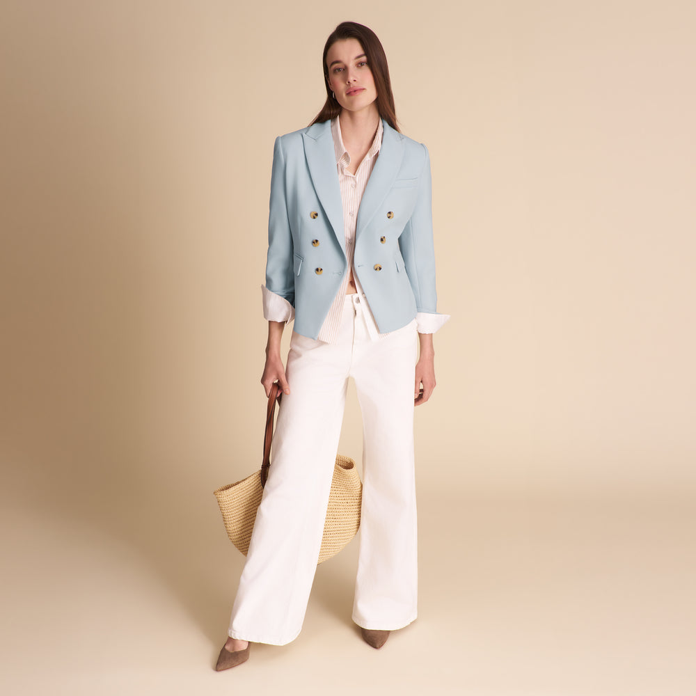 Woman wearing a light blue blazer over a white shirt with white pants, holding a straw bag on a beige background.