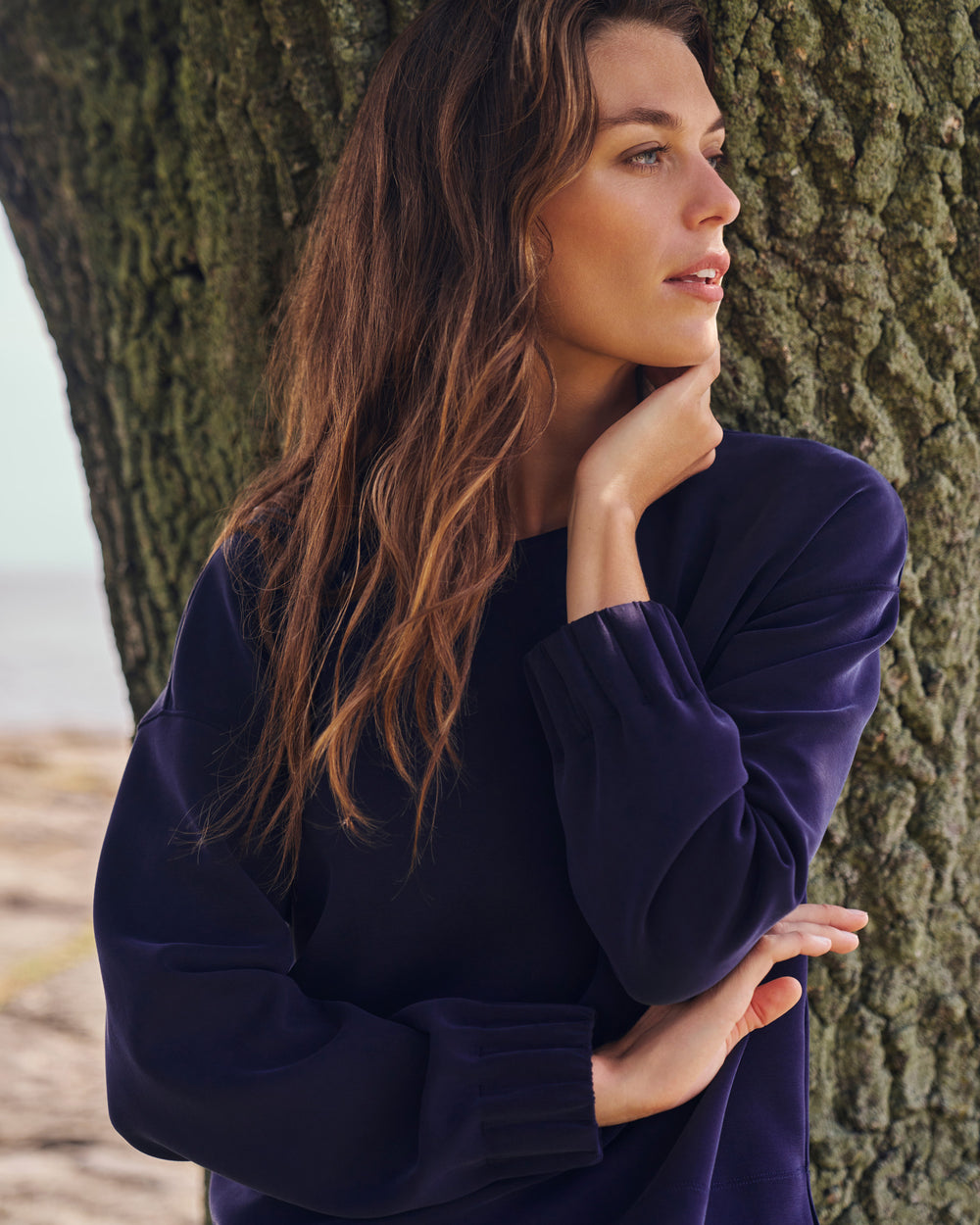 A woman with long brown hair leans against a large tree trunk outdoors, wearing a dark navy long-sleeve top with gathered cuffs.