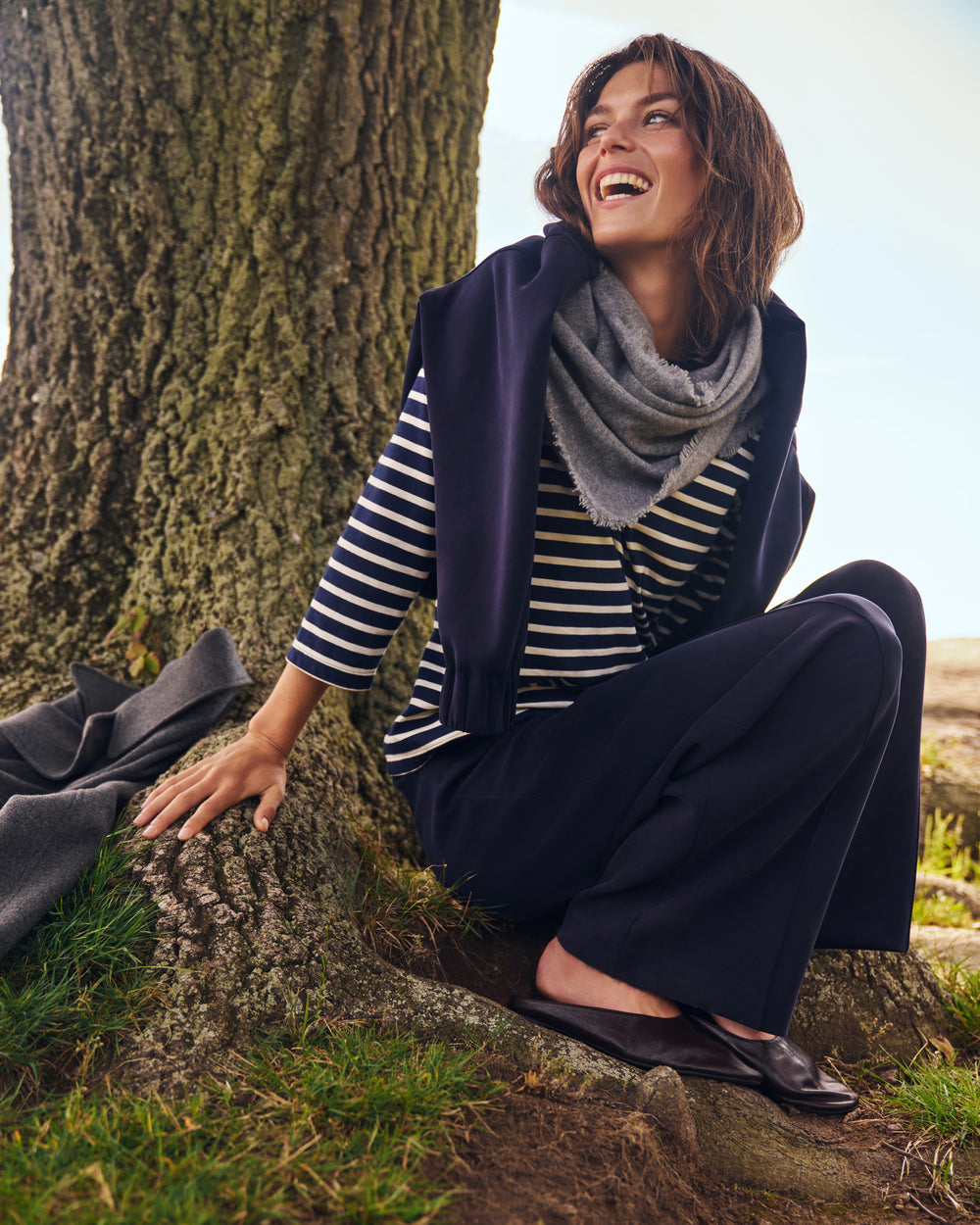 A woman sitting outdoors against a large tree trunk, wearing a navy sleeveless vest, navy wide-leg trousers, a navy and white striped long-sleeve top, a gray knit scarf, and dark flat shoes.