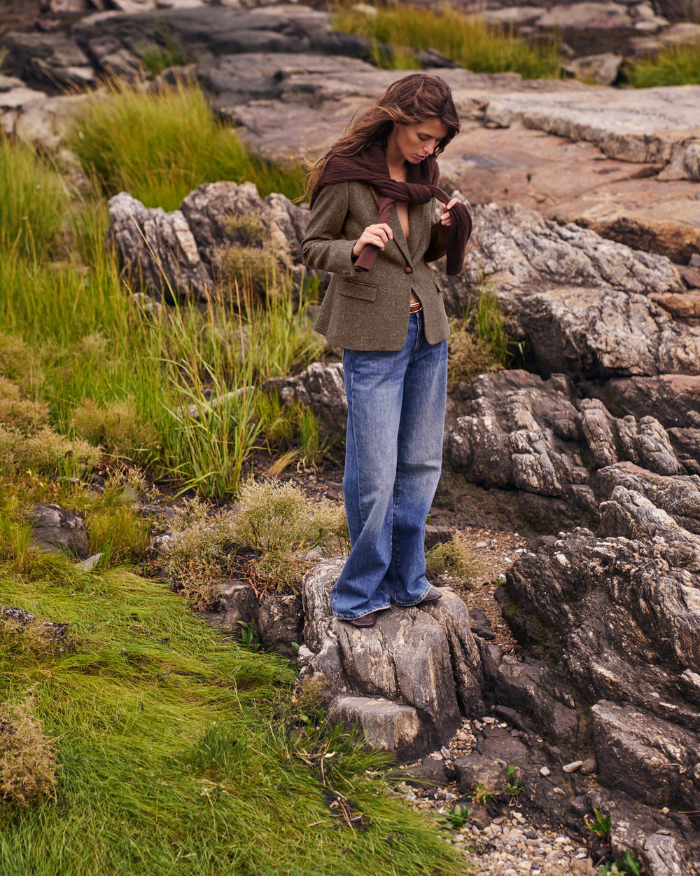 A woman stands on a rock in a rocky, grassy outdoor landscape, wearing an olive green blazer, blue jeans, and a dark brown scarf.