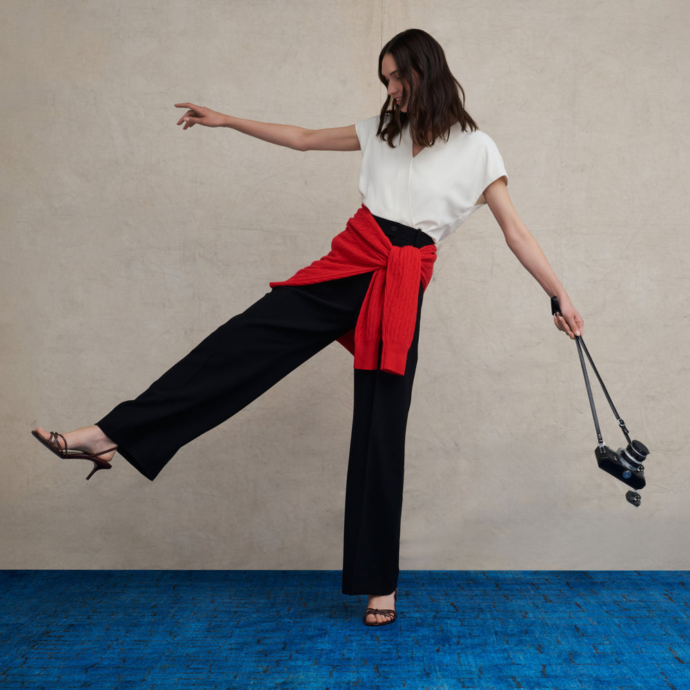 Woman wearing a white v-neck short sleeve top and wide leg black pants on a blue carpet floor and against a beige background