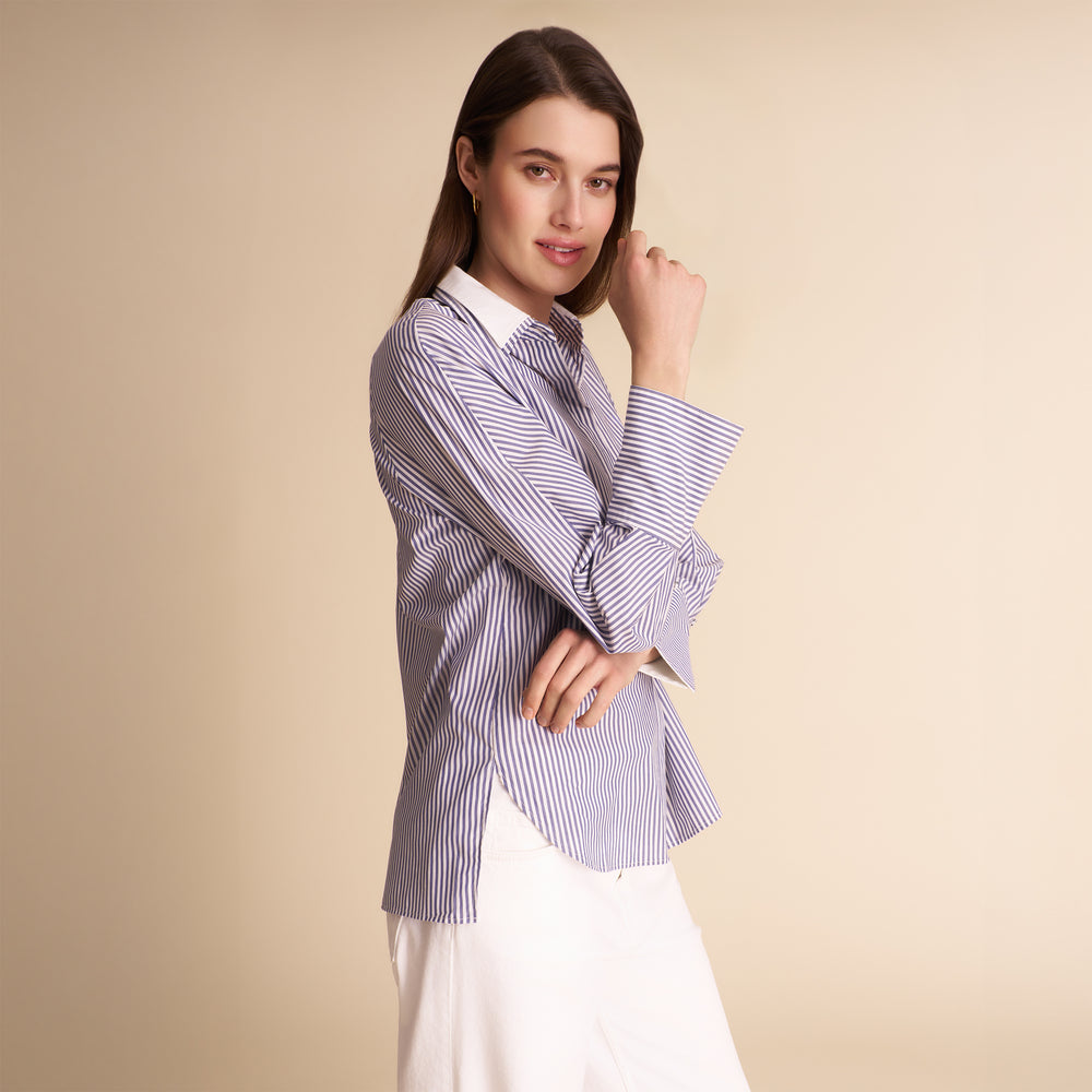 A woman modeling a long-sleeve button-up shirt with thin vertical blue and white stripes, paired with a white skirt, against a solid beige studio background.