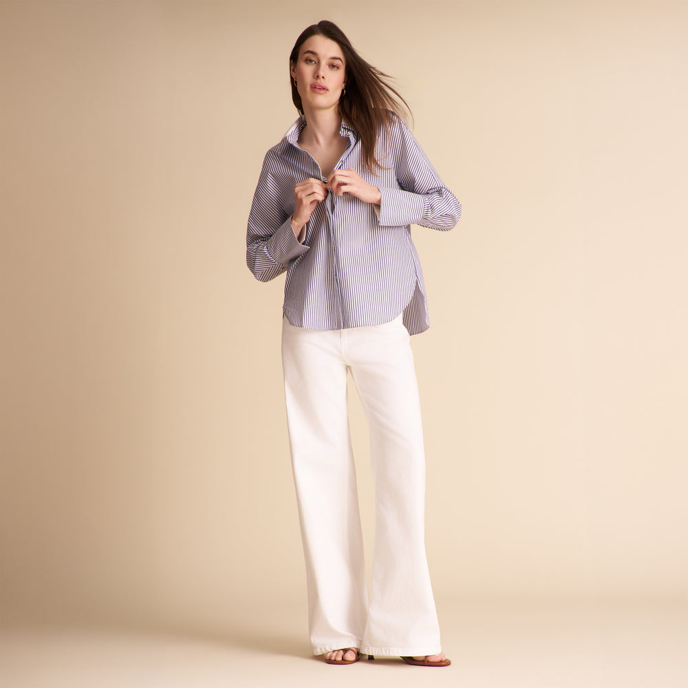 A woman modeling a light blue and white vertically striped button-down shirt with white wide-leg trousers, standing against a solid beige studio background.
