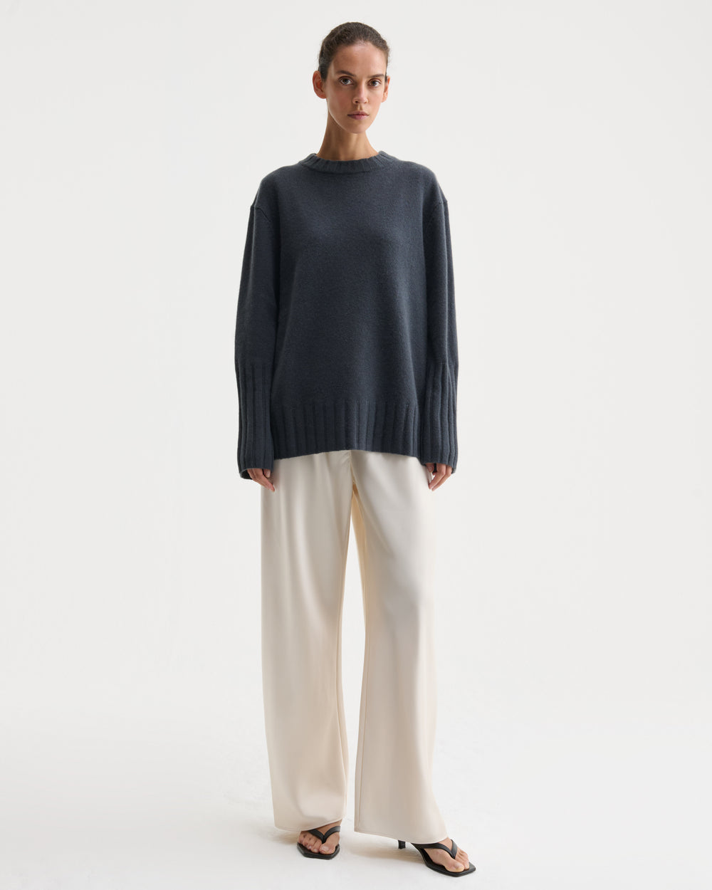 A model stands against a plain light gray studio background wearing an oversized dark charcoal knit sweater, wide-leg cream trousers, and black sandals.