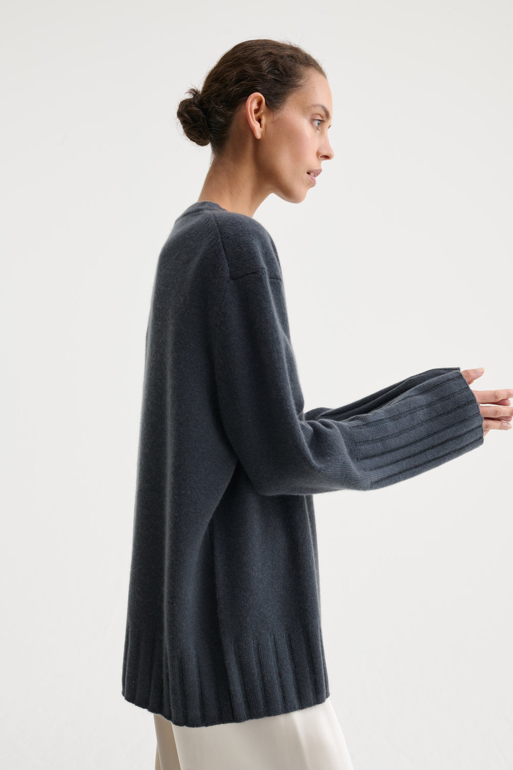 A model stands against a plain light gray studio background wearing an oversized dark charcoal knit sweater, wide-leg cream trousers, and black sandals.