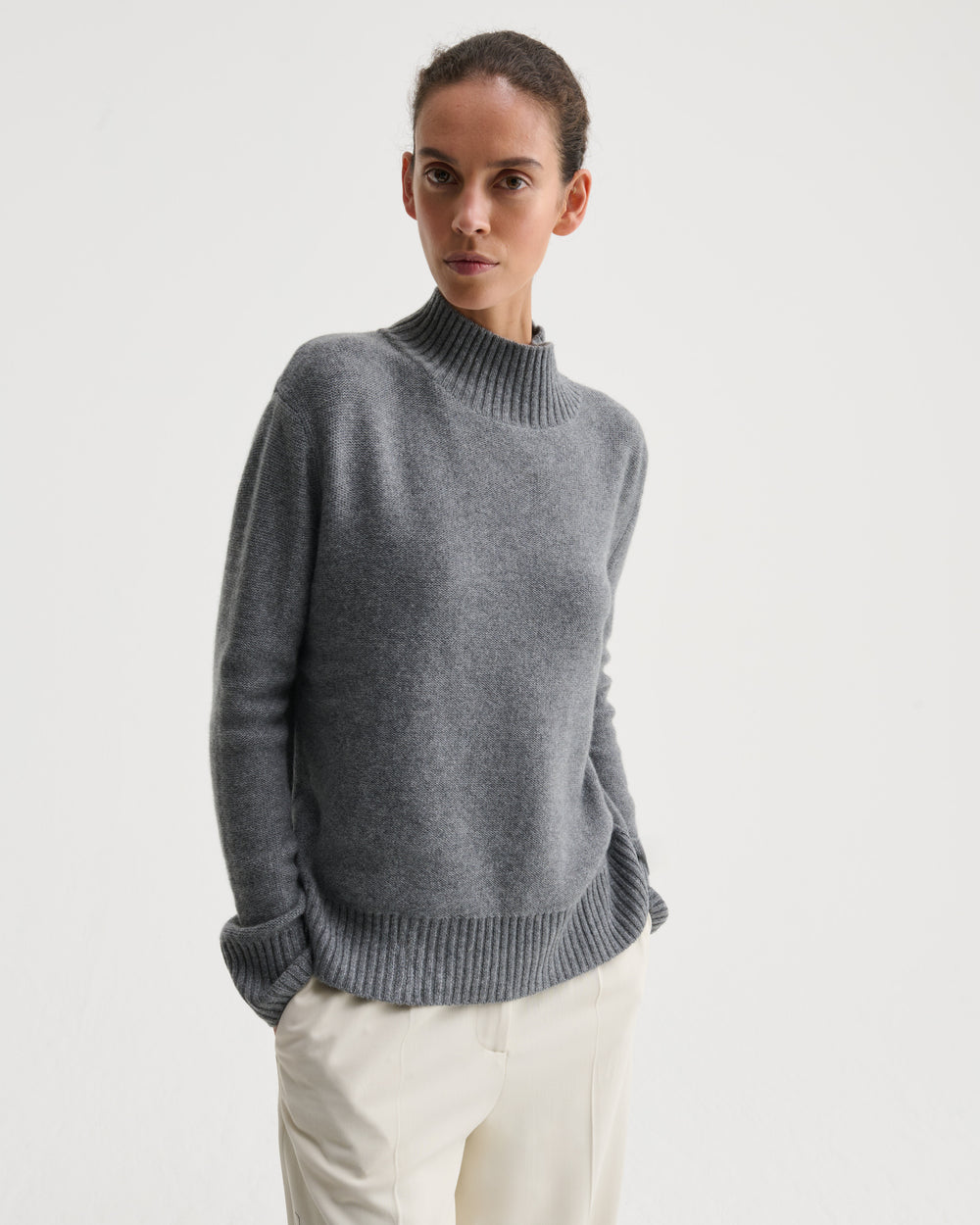 A woman wearing a gray ribbed mock neck sweater and cream trousers, standing against a plain white studio background.