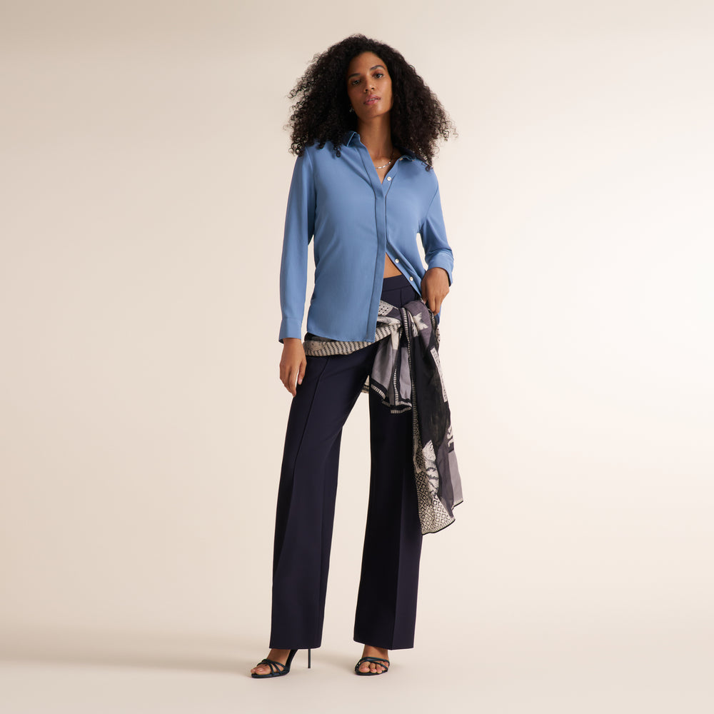 A woman stands against a plain warm beige studio background wearing a light blue button-up shirt, dark navy wide-leg pants, and a patterned gray-and-black scarf tied around her waist.