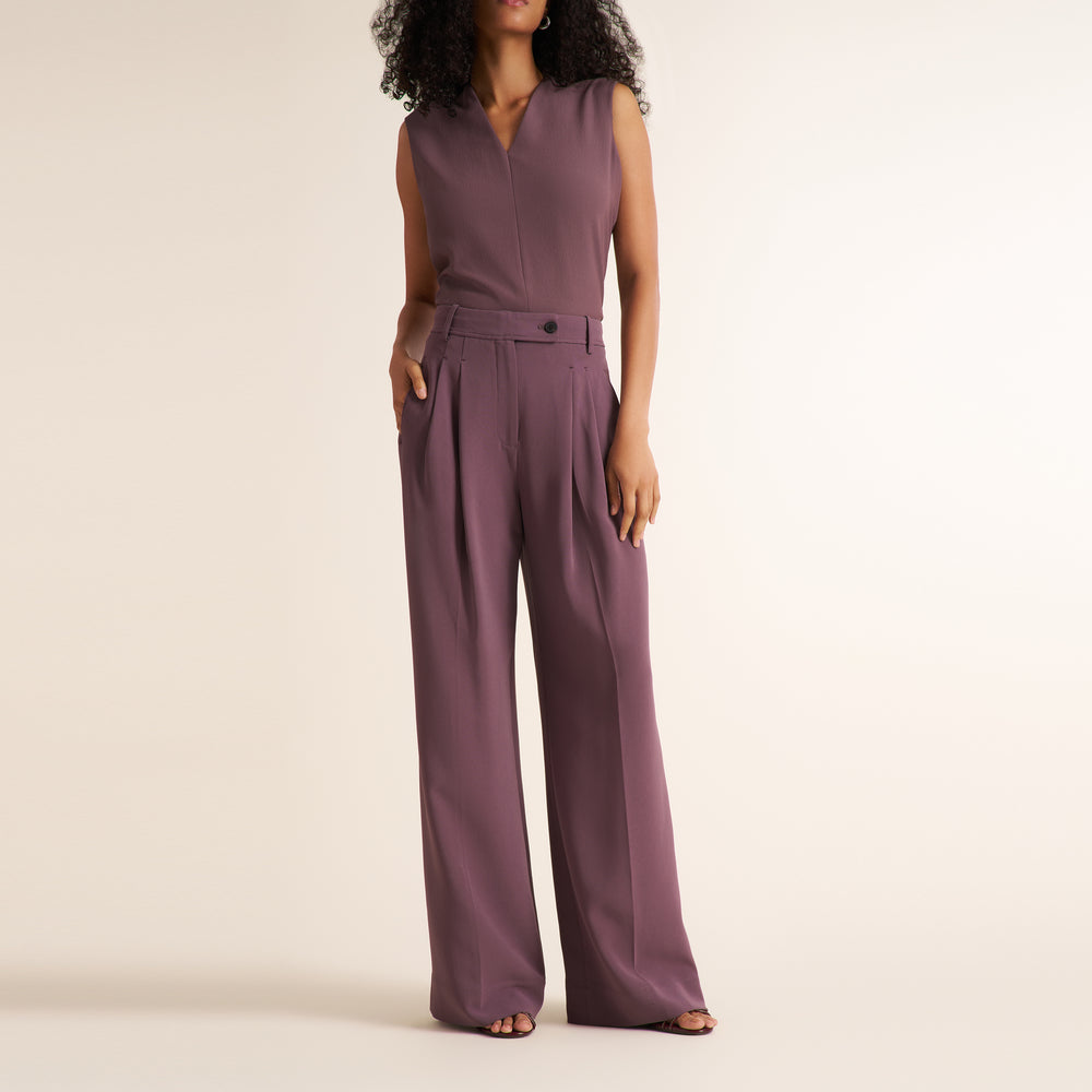 A woman modeling a sleeveless mauve-purple jumpsuit with a V-neckline, front button detail, and wide legs, standing against a plain beige studio background.