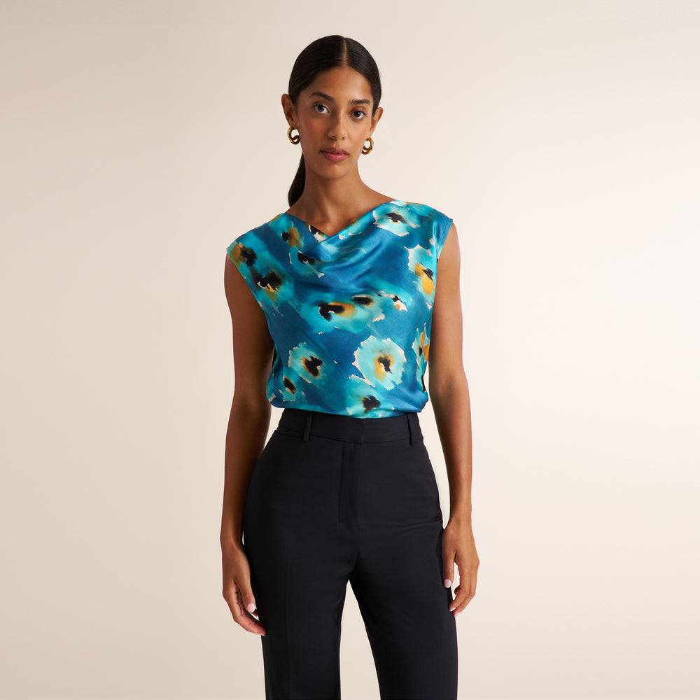 A woman modeling a sleeveless cowl-neck top with a blue floral print, paired with black trousers, against a solid beige background.