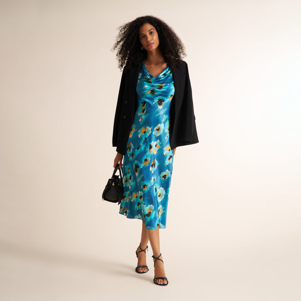 A woman stands against a plain light beige studio background wearing a bright blue floral midi dress with a black blazer, black strappy heels, and carrying a small black handbag.