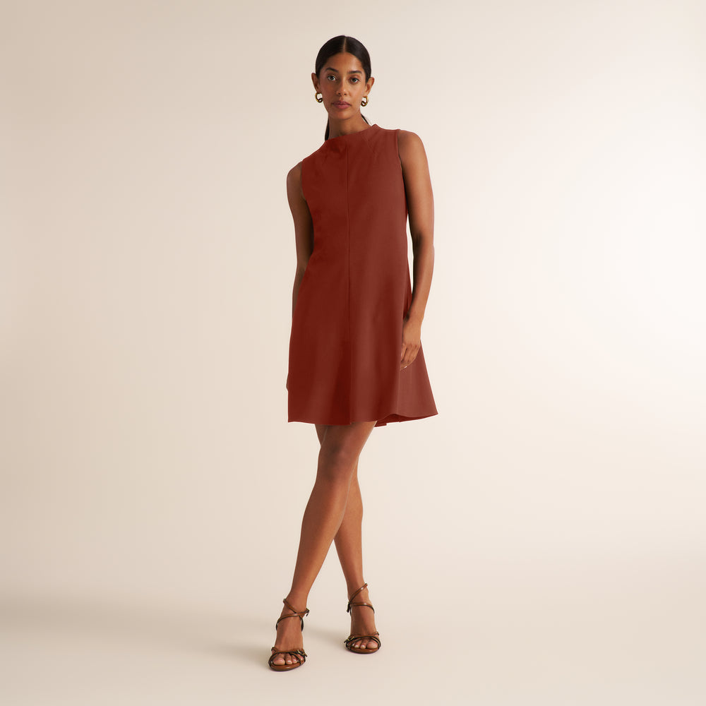 A woman modeling a sleeveless, high-neck, A-line mini dress in a warm rust or terracotta color, paired with brown strappy sandals, against a solid light beige studio background.