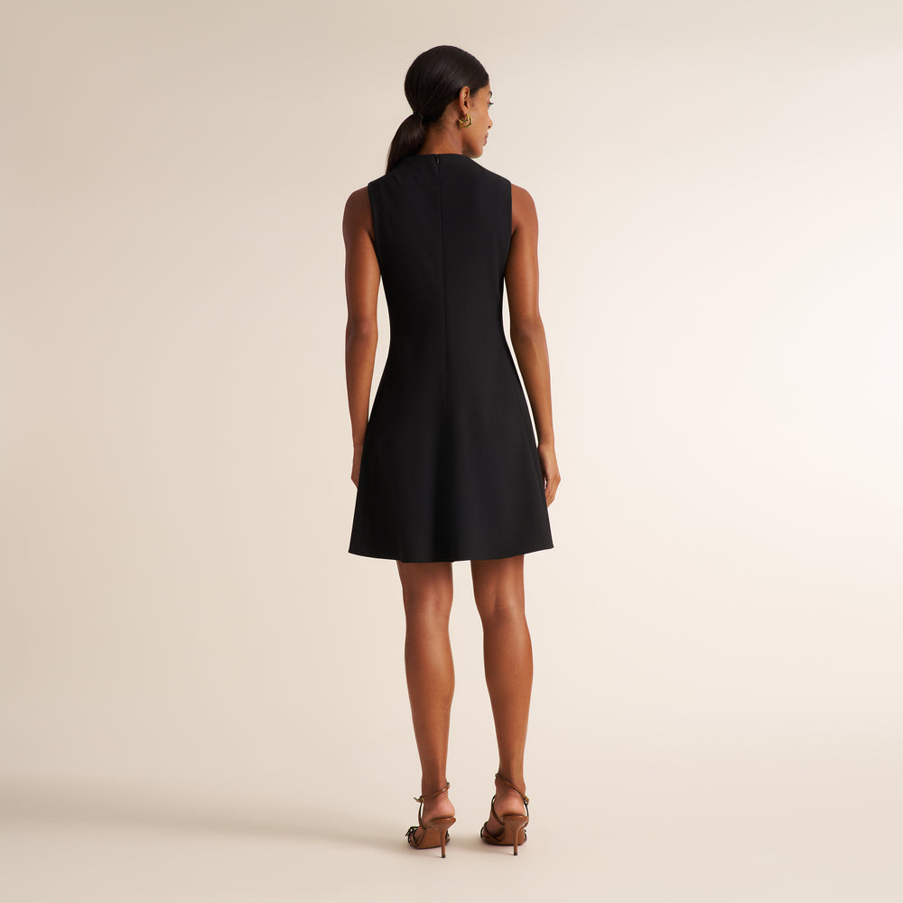 A woman viewed from the back wearing a sleeveless black fit-and-flare dress with a straight neckline and knee-length hem, paired with brown strappy high-heeled sandals against a plain warm beige studio background.
