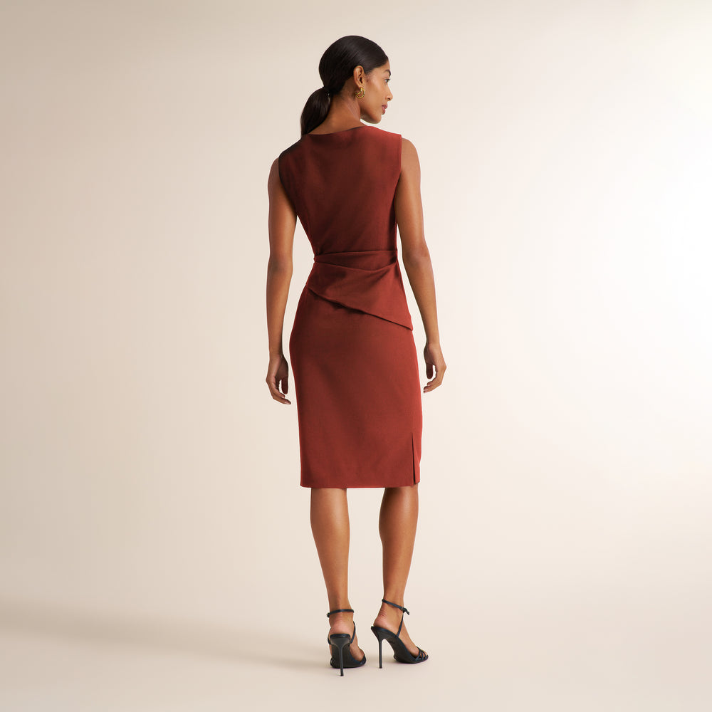 A woman shown from the back wearing a sleeveless rust-brown knee-length dress with a draped wrap-style waist, paired with black ankle-strap heels, against a plain light beige studio background.