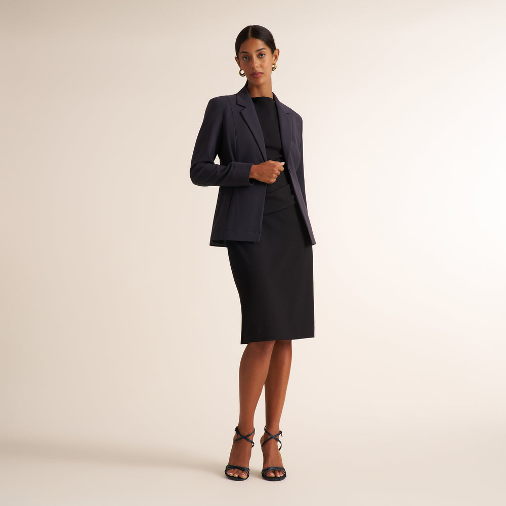 A model stands against a plain light beige studio background wearing a tailored dark navy blazer over a black dress, with black strappy heels.