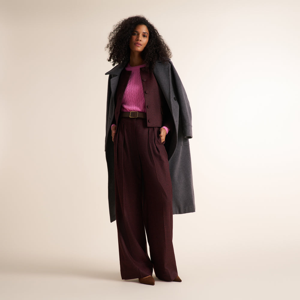 A model stands against a warm beige studio background wearing a dark charcoal overcoat, a deep burgundy vest and matching wide-leg trousers with a brown belt, a bright pink ribbed top, and brown pointed shoes.