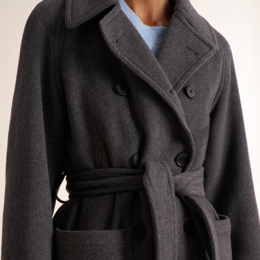 A close-up of a person wearing a dark charcoal gray double-breasted wool coat with a matching waist tie belt, layered over a light blue top.