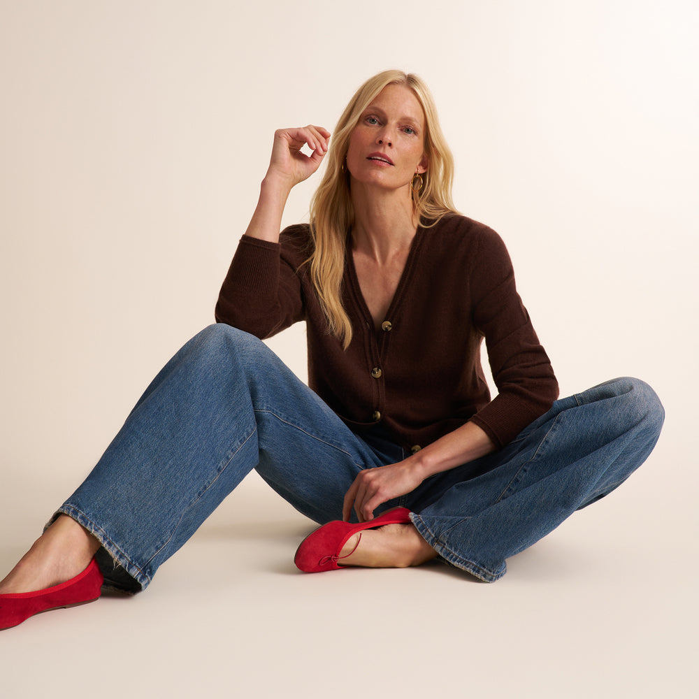 A blonde woman sits cross-legged on a warm off-white studio floor, wearing a dark brown button-front cardigan, wide-leg blue jeans, and bright red flats.